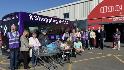 A purple minibus with white Shopping UnLtd branding on it. It is parked outside the door of the supermarket Alliance. Standing and in wheelchairs alongside the minibus with empty trolleys are a variety of people using the service wearing sunflower lanyards. Volunteers are wearing purple tabbards.