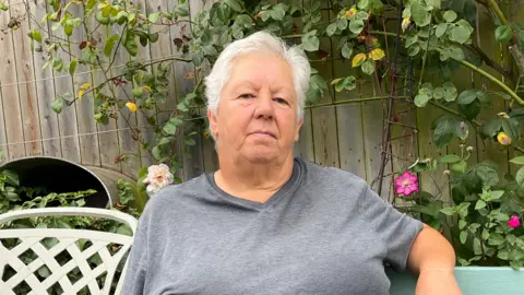 Judith Quérée is sitting on a bench in a garden. Plants are visible behind her. She has short white hair and is wearing a grey T-shirt. She has a muted smile.