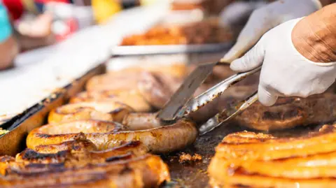 Getty Images A cook grills a number of sausages on a cooker. The are browned and slightly charred, the cook wears white gloves and is holding tongs