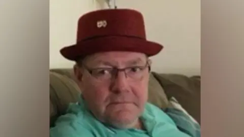 A grainy photograph of Gary Piatek, a white, middle-aged man wearing a red hat and a green polo shirt. He is sitting on a sofa. 