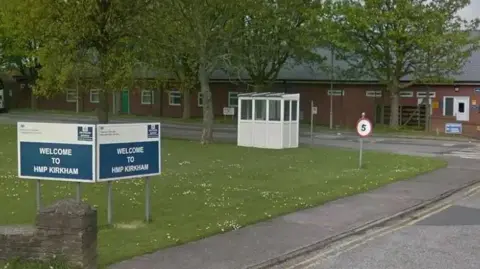 Entrance to HMP Kirkham prison building showing a sign which reads: "Welcome to HMP Kirkham". It is on grass which has a path up the side a 5mph sign and a white shelter if you turned left.