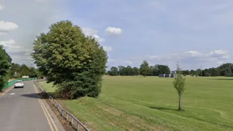 Google A general view of a large playing field, which has a narrow road running next to it.