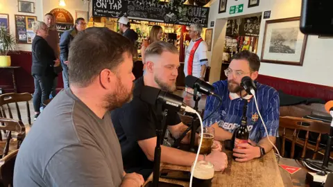 Sam Read/BBC Three men sit around a microphone on a pub table, with drinks. They are in a pub and people are milling at the bar.