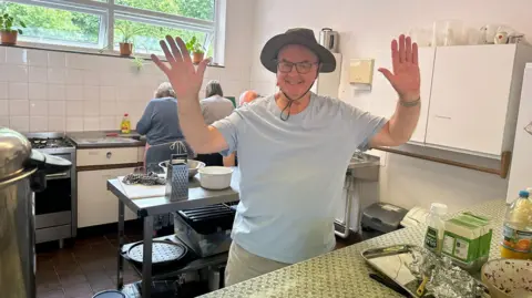 Cad Taylor, BBC A man in a hat stands in a kitchen with his arms in the air