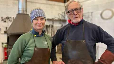 BBC Melissa Cole (left) and Hector Cole (right). Melissa has blonde hair that is held back by a blue fabric hairband. She is wearing a blue shirt, green jumper with a brown leather apron. Hector is wearing a navy blue top with a red bandanna tied as a scarf and a brown leather apron. He is also wearing a pair of dark rimmed glasses.