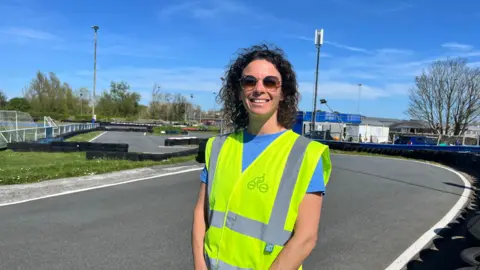BBC A lady in a high viz yellow jacket stands on a tarmac road. She's wearing sunglasses with a dark blue sky in the background. 