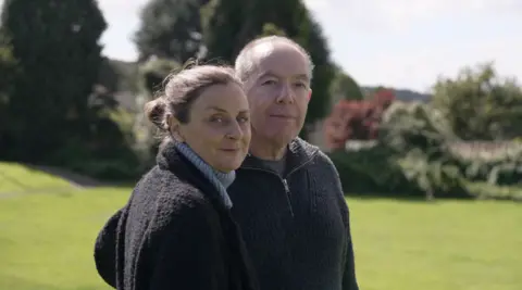 Andy Alcroft/BBC Portrait of Fergus' parents standing in the park where the sapling is being planted.