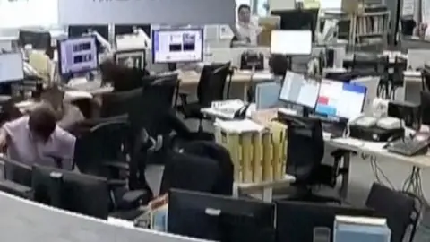 A man in a shirt ducks beneath a desk, surrounded by shaking office furniture and screens.