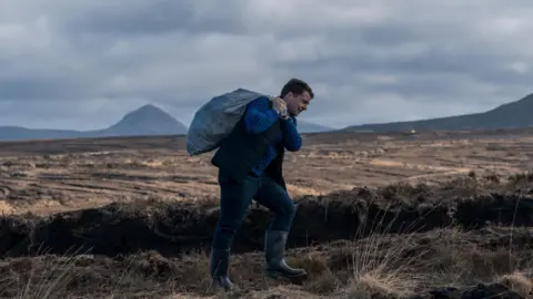 Wearing a blue turtleneck, dark vest, trousers and wellie boots, Alex Murphy carries a heavy sack over his shoulder as he walks through an empty marshland.