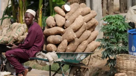 A yam seller dressed in his traditional attire with a cap sits on his wheel barrow containing piles of yam