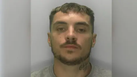 A police custody image of Tyler Doyle. He has short brown hair, a moustache and a beard. He has a tattoo on the left side of his neck.