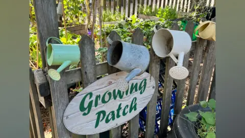 Beach Babies A close up shot of four small metal watering cans hanging on a fence post which has an oval sign saying Growing Patch. Behind are plants and flowers growing in raised beds.