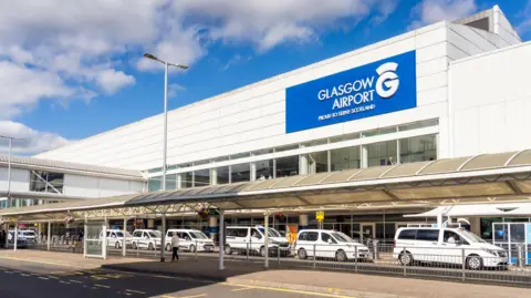 The exterior of Glasgow Airport's main building. It is a large white rectangle shaped structure with a blue sign on a wall that says: "Glasgow Airport. Proud to serve Scotland". A row of white taxis are parked outside. In the foreground is a long bus shelter.