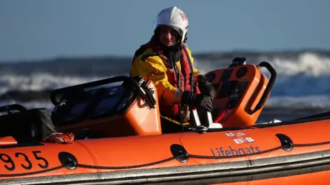 Helen Cowan Carl steers an orange lifeboat through waves on the sea. He is wearing a white helmet, yellow dry suit and red vest.