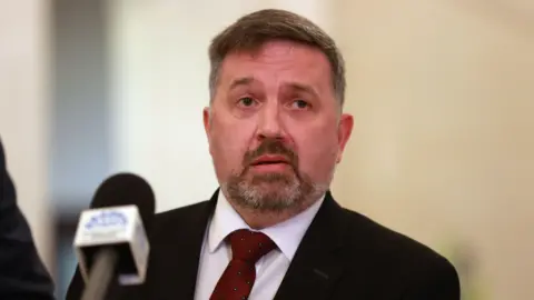 Getty Images Robin Swann is standing in front of a mic that says Northern Ireland Assembly. He's wearing a black suit with a white shirt and a maroon tie. 