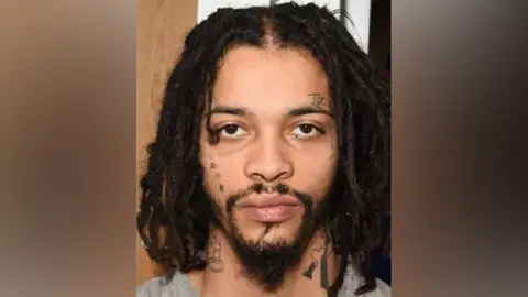 Met Police Zak Baako has long black hair, face tattoos and a goatee. 