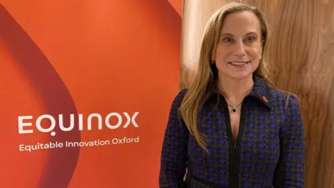 A smartly dressed woman is smiling at the camera. Behind her is a banner with the words EQUINOX, Equitable Innovation Oxford.