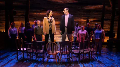 Craig Sugden A production shot from a scene in Come From Away. A man and a woman have their heads turned towards one another as they are stood on a line of wooden chairs. A group of people stand behind them, looking at them. The man is dressed in a blazer and chinos, and the woman in black trousers and a sand-coloured jacket. The spotlight is on the couple. 