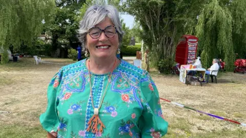 A woman wearing a bright green and blue top with a brown necklace. She has short grey hair and is wearing glasses. Behind her are trees and a red phone box. The sun is shining.