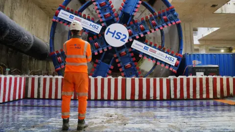 PA Media A man in fluro orange stands in front of a boring machine with HS2 written on it.