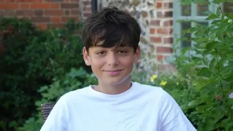Kaan a boy with brunette hair who is sitting outside in front of a bush. He is wearing a white T-shirt and is looking directly at the camera and smiling.