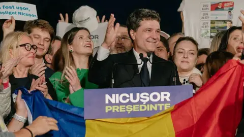 Andrei Pungovschi/Getty Images A smiling man in a suit stands with supporters in front of a Romanian flag and a lectern that says Nicusor president
