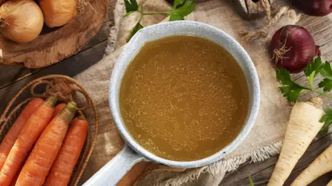 Getty Images Chicken bone broth or soup in a saucepan with fresh vegetables on a wooden table, top view