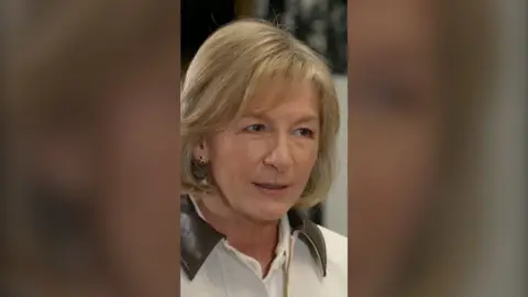 A woman with shoulder length blonde hair, wearing a white jacket with a brown collar