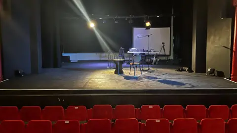 Rows of red seats in a theatre sit in front of a black stage which has a table and chairs along with theatre equipment