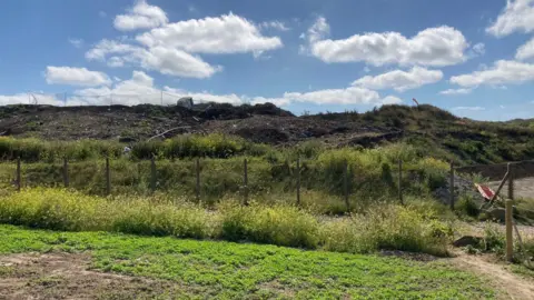 A landfill site showing a mound of dirt littered with rubbish including a boat. It is a partly cloudy day.