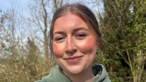 Rosie is a young woman smiling to the camera, she is wearing a green hoodie and has light brown hair, she is standing in front of trees. 
