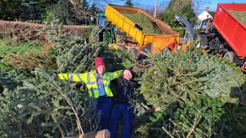 St Margaret's Hospice Two people lying outside on a pile of Christmas trees, with lorries in the background full of wood chippings. 