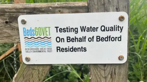 BedsGOVET A close up of a sign on a gate. It tells people that testing of water quality by the Bedfordshire Great Ouse Valley Environmental Trust is taking place.