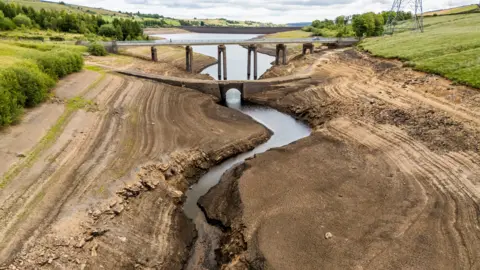 Danny Lawson/ PA Wire A dried-up reservoir in a rural area reveals cracked earth and sediment layers, with a narrow stream winding through the centre. The stream passes under an old stone bridge and a modern concrete bridge. Surrounding the exposed terrain are grassy fields, scattered trees, and a power line tower.