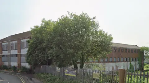 Google The same factory seen from further away, showing its full size. A long brick warehouse is behind the main road, with trees and grass around it. There are many windows, some boarded up. Despite being derelict it looks neat.
