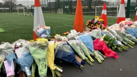 Dozens of bouquets of flowers placed at the side of a football pitch, many with handwritten messages in memory of Poppy.  