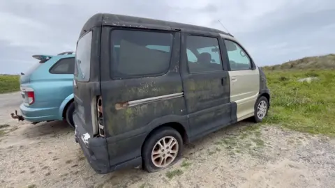 BBC A weathered and neglected black van with moss and rust on the body panels, missing tail lights and a flat rear tyre parked on a dirt car park near grassy dunes. The sky above is a leaden grey colour with a blue SUV parked behind the van. 