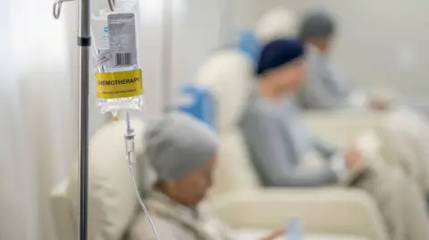 Getty Images An IV bag with clear liquid in it with a yellow sticker saying "chemotherapy". Three patients can be seen sitting in chairs behind it.