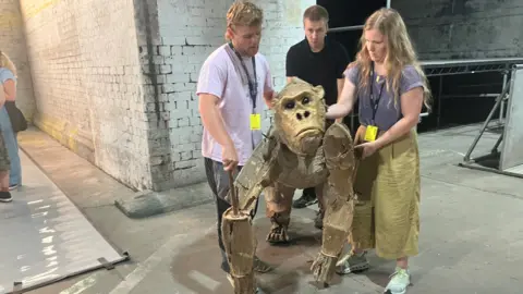 Photograph of three people controlling a puppet gorilla. The picture was taken inside a rehearsal space under railway arches in Manchester city centre.