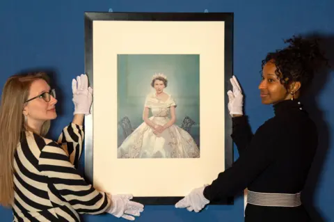 Jane Barlow/PA Wire Two curators hold a picture of a picure of Princess Elizabeth who is wearing a white dress. 