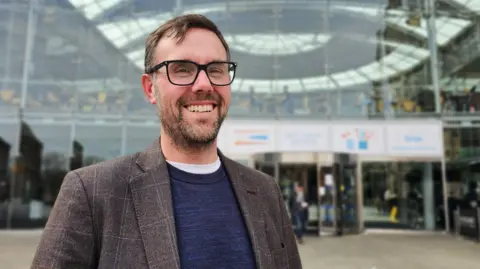 Paul Moseley/BBC James Wright is wearing a brown suit jacket over a blue jumper and white t-shirt. He is standing outside the glass-fronted Forum building in Norwich.