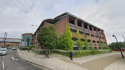 The four-storey St Mary's multi-story car park clad in red bricks with multiple large gaps in the wall. The sky is grey. A road runs to the left of it with cars coming towards the camera. A few people are walking on the street. In the distance is a covered bridge over the road with the worlds Sunderland City Centre written out across its windows.