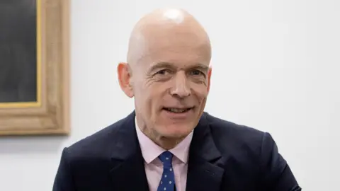 PA Media Sir Adrian Fulford, who has a bald head and is wearing a navy blue suit over a pink collared shirt, smiles at the camera