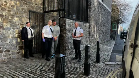 Paul Butler, 53, of Stangray Avenue, Plymouth, being escorted out of Plymouth Magistrates Court on Monday 27 January. He is wearing a grey tracksuit and blue flip flops. He is being escorted by three prison officers.