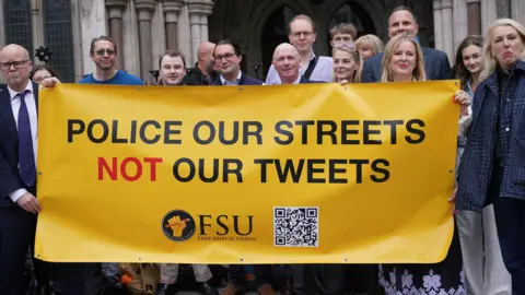 A group of people including men and women hold up a yellow banner with the wording "POLICE OUR STREETS NOT OUR TWEETS" in black lettering. There is an FSU (Free Speech Union) logo and a QR code on the banner.
