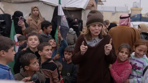 Kabawat in a brown jumper and woollen hat, surrounded by about 10 children, several of whom are looking up at her. Some are smiling. She is speaking and gesturing with her two forefingers outstretched. Tents in the camp for displaced people can be seen behind her. 