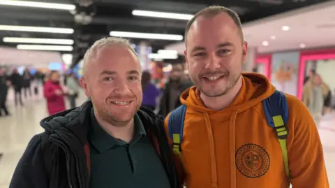 Craig Ballentine pictured in a camel coloured hooded top with his friend Sean who is wearing a dark top and jacket at the airport. They are smiling at the camera