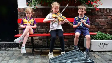 Three children are sitting on a red painted wooden bench on a cobbled street. A boy and girl are playing the tin whistle and another girl is playing the violin. 