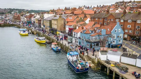 Getty Images A harbour with small fishing boats moored along the waterfront, backed by colourful buildings with red‑tiled roofs. People walk along the quay, with houses rising up the hillside.