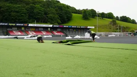 The image shows a football stadium undergoing resurfacing work. In the foreground there is a section of green artificial turf, with two workers handling a rolled section. A long strip of turf is midway through being unrolled. Behind the workers is the main playing surface area. White bundles are scattered around the edges. To the right of the image a machinery arm is visible.